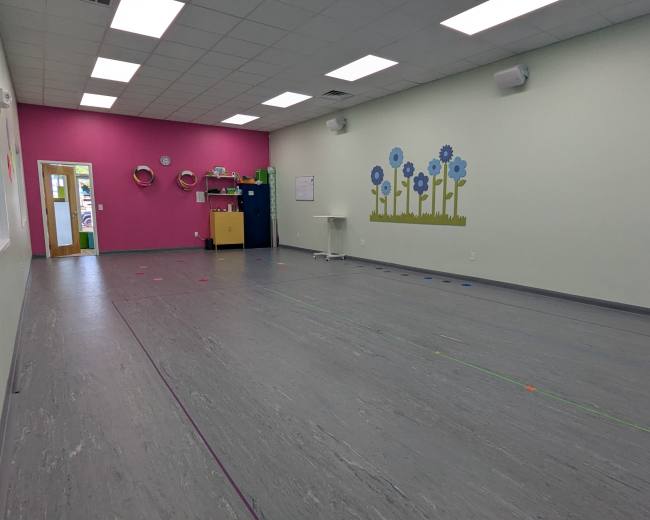 The image shows a brightly colored classroom with a pink and light green wall, featuring a flower mural, and an open space with a table and storage units along the walls.