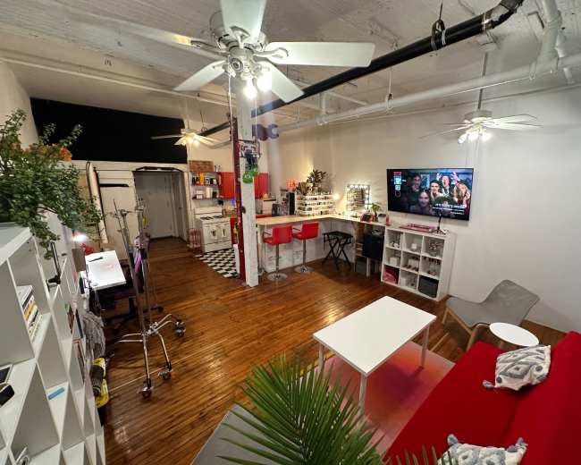 The image shows a spacious room with wooden flooring, featuring a red sofa, a white coffee table, a kitchen area in the background, and a television mounted on the wall.