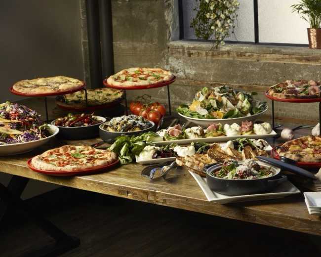 A table is set with various types of pizzas, salads, and side dishes arranged on multiple platters against a backdrop of large windows and potted plants.