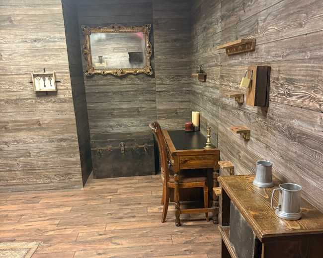 The image shows a dimly lit room with wooden walls and flooring, featuring a desk with a chair, a mirror, shelves, and a cabinet.