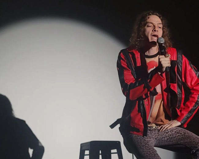 A performer in a red jacket and leopard print pants sits on a stool while speaking into a microphone against a lit backdrop.