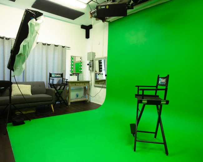 The image shows a green screen setup in a studio with a director's chair, lighting equipment, a couch, and mirrors on the wall.