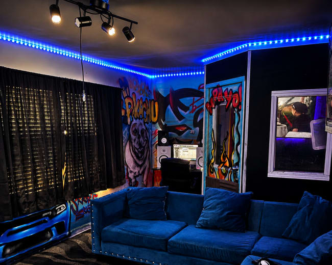 The image shows a vibrant room featuring a blue sectional couch, graffiti-covered walls, a desk with a computer, and a large window covered by black curtains.