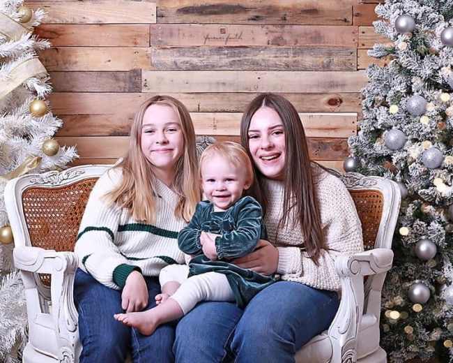 Two girls sit on a sofa beside a toddler, with decorated Christmas trees in the background.