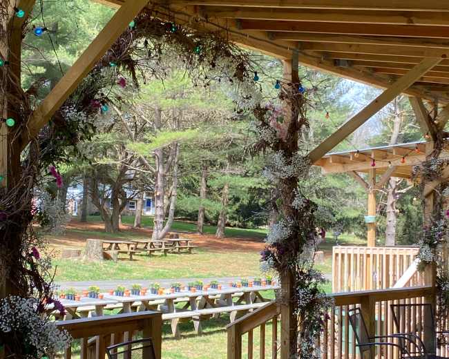 A wooden porch with black chairs overlooks a grassy area with picnic tables and flower planters in the background.