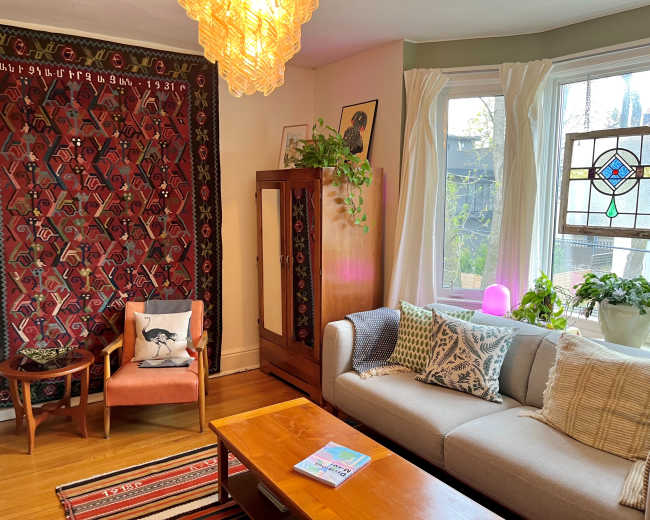 The scene depicts a cozy living room with a patterned wall tapestry, a light fixture hanging from the ceiling, a sofa adorned with decorative pillows, and a mix of plants near a large window.