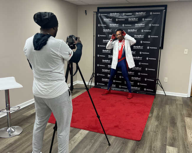 A person in a lab coat poses for a photo in front of a backdrop while another person operates a camera.