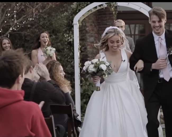 A bride and groom walk down the aisle together, smiling, as guests cheer and applaud in a garden setting.