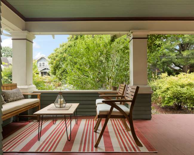 The image shows a porch with a seating area featuring two chairs and a coffee table, surrounded by greenery and a striped rug.
