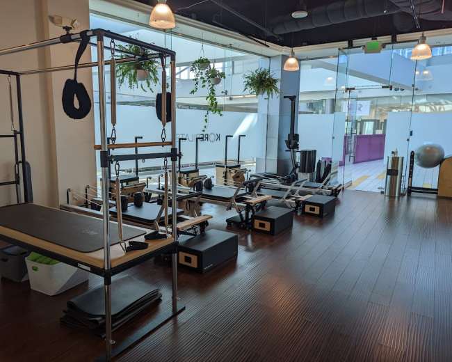 The image shows a brightly lit fitness studio featuring various Pilates equipment, including reformers, a reformer barrel, and mats arranged neatly on the wooden floor.