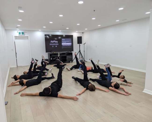 A group of individuals is practicing a coordinated floor exercise in a well-lit room with a screen displaying an AirPlay connection.