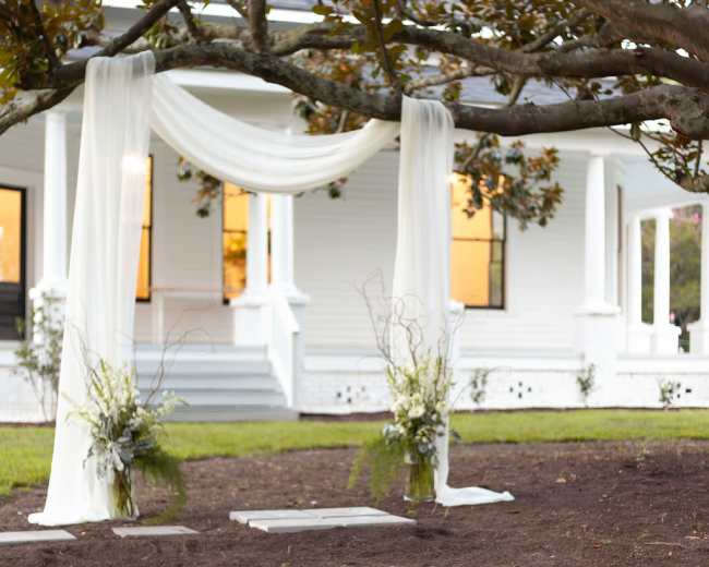 An outdoor wedding setup features a decorative draped arch under a large tree, with white chairs arranged in front on a grassy area.