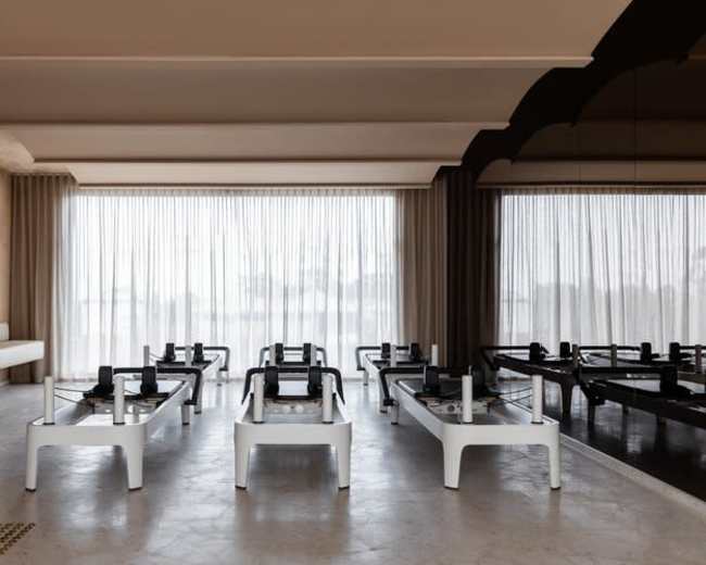 The image depicts a modern fitness studio with several reformer machines arranged in rows, large windows allowing natural light to flood the space, and a neutral color palette.