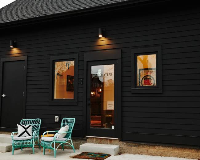 A black wooden house exterior featuring a main entrance door and two windows, with green wicker chairs placed outside on a concrete walkway.