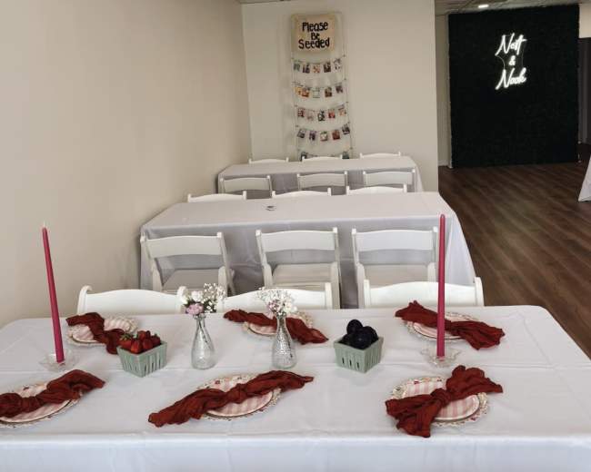 The image shows a dining area with white tables set for a meal, featuring red candles, floral arrangements, and napkins.