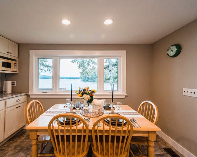 A dining area features a wooden table set for a meal, with a bouquet of flowers, and large windows overlooking a lake.