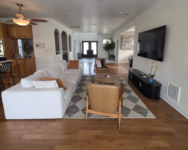 The image shows a modern living room with a white sectional sofa, two wooden chairs, a coffee table, and a large wall-mounted television, all arranged on a patterned rug over hardwood flooring.