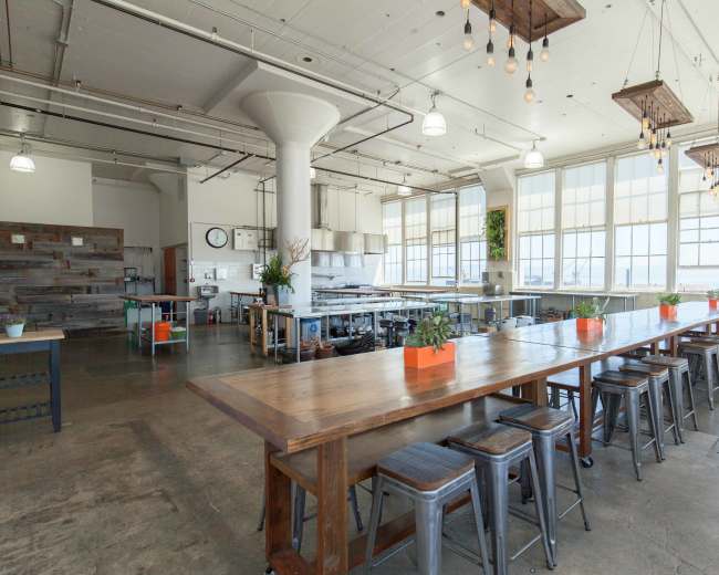 A spacious industrial-style kitchen and dining area with large windows, a long wooden table, and metal stools.