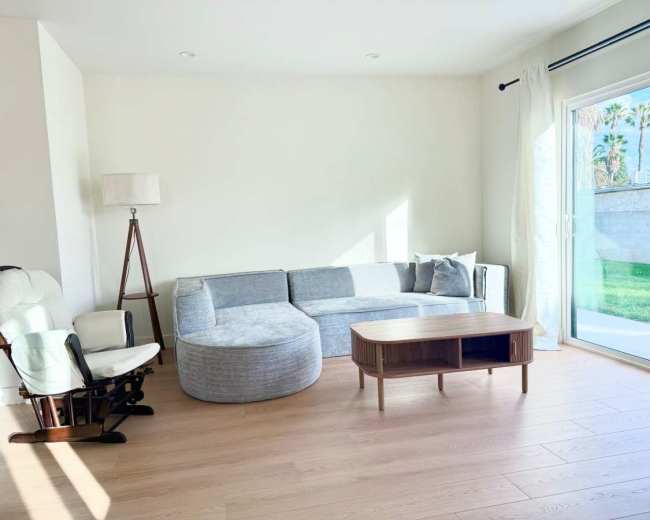 The image shows a modern living room with a gray sectional sofa, a round ottoman, a wooden coffee table, and a white rocking chair beside a large window.