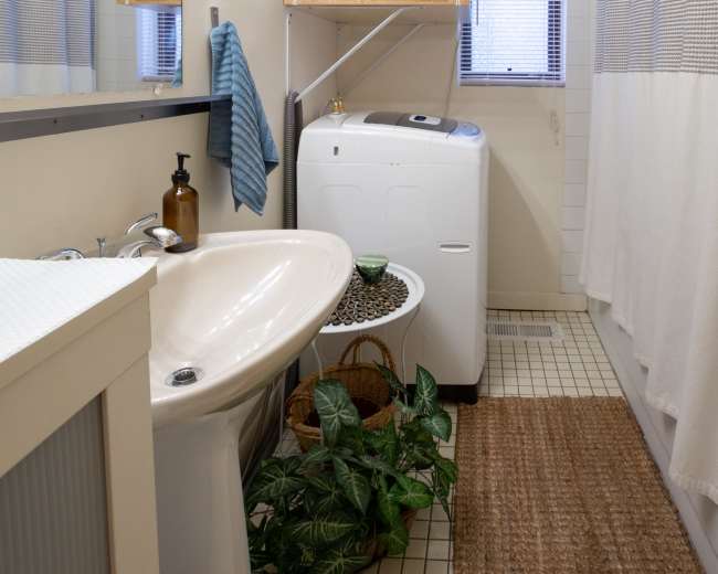 The image shows a small laundry area with a white washing machine and dryer stacked above, beside a sink and a mirror, all set in a bathroom with light-colored walls and a woven rug.