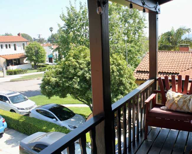 The image shows a balcony view overlooking a street with parked cars and houses, featuring a red adirondack chair and green trees in the foreground.