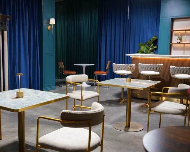 The image shows a stylish indoor space featuring modern tables and chairs, a bar area, and blue curtains.