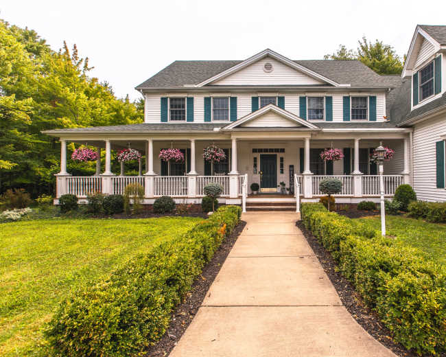 A large, two-story house with a front porch and hanging flower baskets is surrounded by a neatly landscaped yard and pathway.