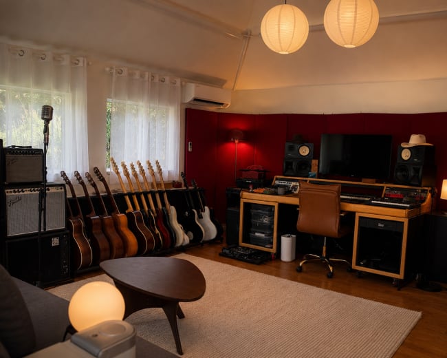 A home recording studio features a row of electric and acoustic guitars against the wall, a mixing desk with monitors, and soft lighting from paper lanterns.