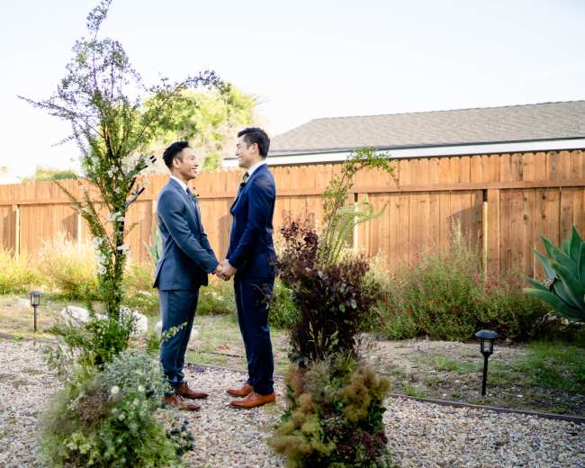Two men in suits stand facing each other, holding hands amidst a garden with greenery and a wooden fence in the background.