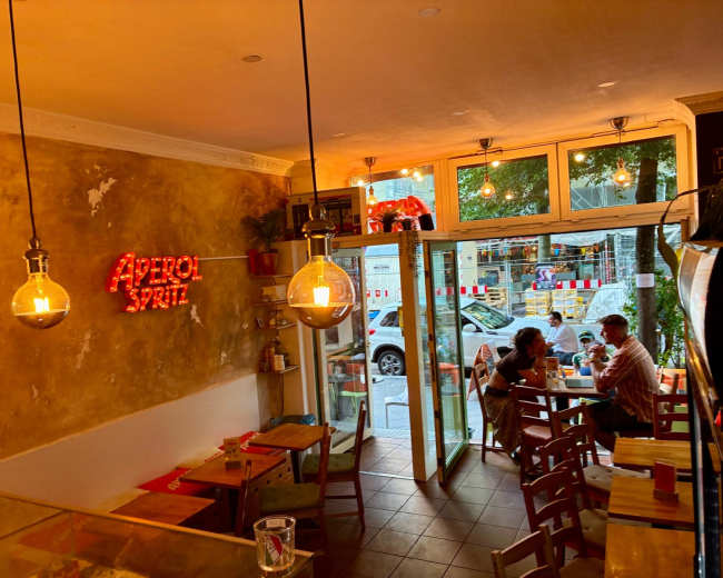 The image shows a cozy café interior with warm lighting, featuring a neon sign that reads "Aperol Spritz," and two patrons seated at a table near a large glass door.