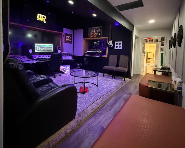 The image shows a recording studio with a control desk, comfortable seating, and soundproofing panels on the walls.