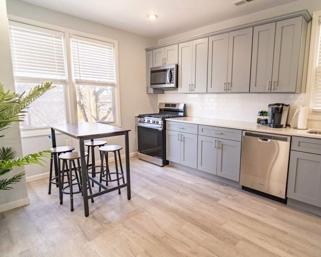A modern kitchen with light gray cabinetry, stainless steel appliances, and a small dining table with four black stools.