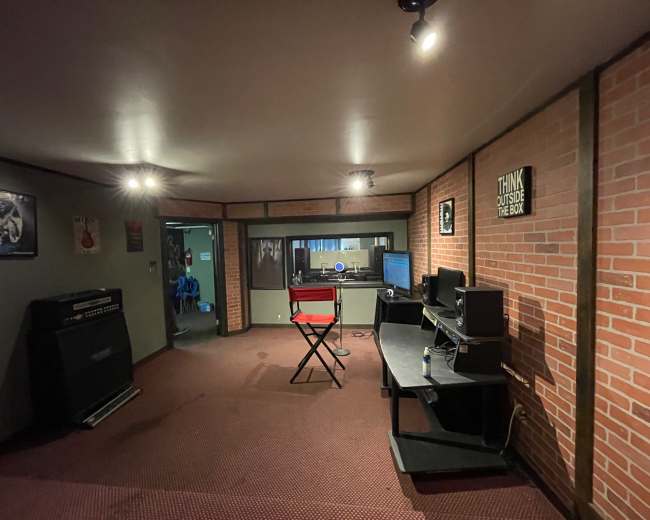 The image shows a music studio with brick walls, a black chair, a desk with a computer, and amplifiers in the corner.