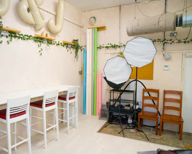 The image shows a studio space with a white bar table and stools, decorated with plants, and featuring professional lighting equipment and colorful backdrops against a light-colored wall.