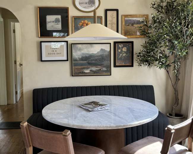 The image shows a dining area with a round marble table, two brown chairs, and a gallery wall featuring various framed artworks.