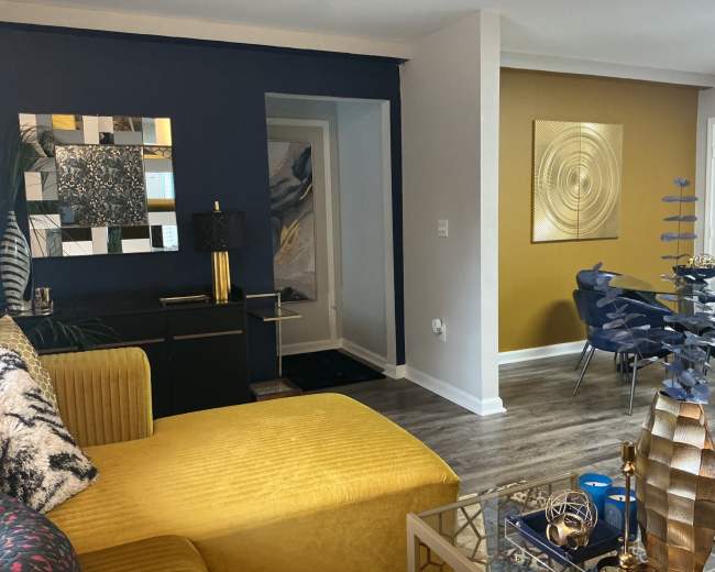 The image shows a modern living space featuring a yellow sofa, an abstract wall art piece, and a dining area with a round table and blue chairs.