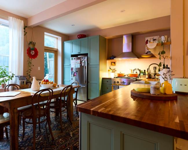 A bright kitchen features a wooden dining table surrounded by chairs, teal cabinets, and a stainless steel refrigerator, with sunlight streaming through a large window.
