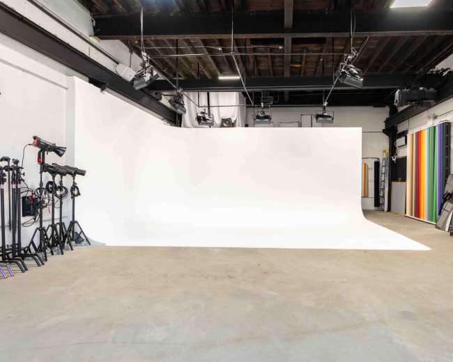 The image shows a spacious photography studio with a large white backdrop, various lighting equipment, and a colorful striped wall on one side.