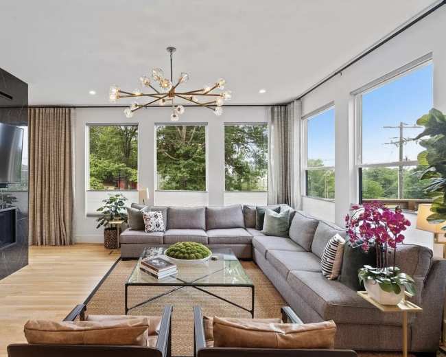 The image shows a modern living room with a gray sectional sofa, a coffee table, large windows, and decorative plants.