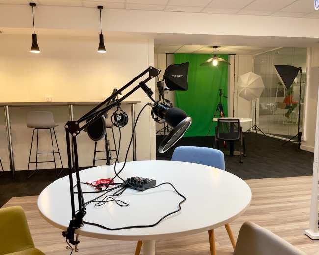 The image shows a studio setup with a round white table in the foreground, featuring a microphone and headphones, and a green screen along with lighting equipment in the background.
