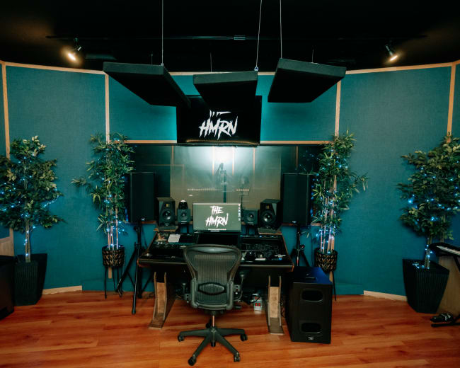 The image shows a music production studio with a central desk, computers, speakers, acoustic panels on the walls, and potted plants.