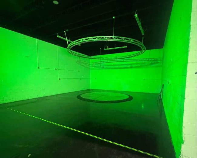 The image shows a dimly lit space with green walls, a circular floor area, and a suspended circular truss overhead.