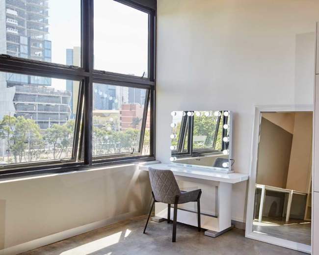 A modern vanity area with a lighted mirror and a chair is located beside large windows in a room with a view of a city skyline.