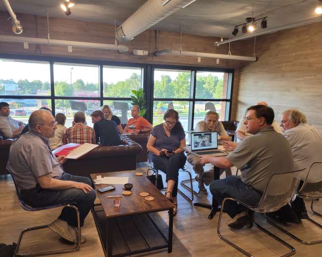 A group of people is gathered in a well-lit café, engaged in conversation and using laptops around wooden tables.