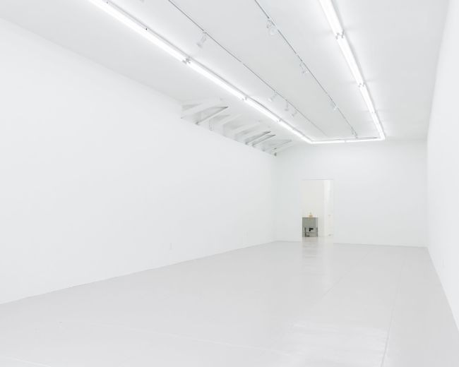 The image shows a minimally decorated white room with bright overhead lighting and a single door.