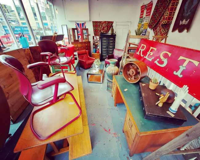 The image shows a cozy vintage shop filled with various pieces of furniture, including chairs, tables, and decorative items, arranged against a backdrop of patterned textiles and wall art.