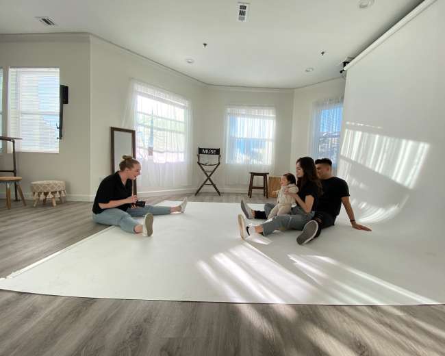A group of four people sits on a white floor in a bright studio with large windows and a backdrop.