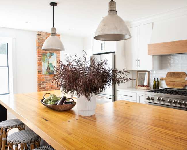 A modern kitchen features a large wooden island with a potted plant at the center, flanked by two pendant lights and surrounded by white cabinets and exposed brick walls.