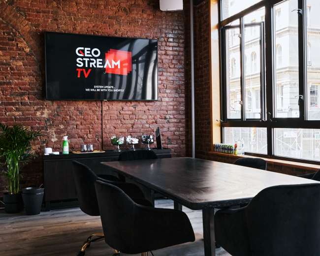 A sleek conference room features a large black table surrounded by chairs, with a television displaying the logo for "CEO Stream TV" on a brick wall.
