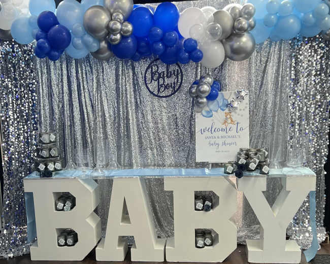 A decorated backdrop for a baby shower, featuring large white letters spelling "BABY," colorful balloons, and a welcome sign.